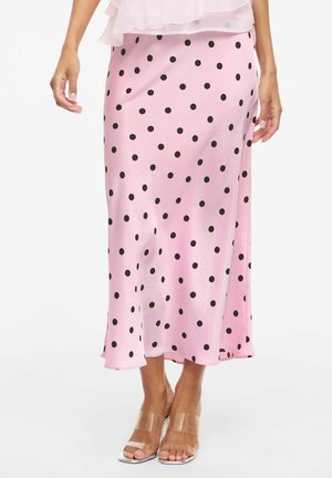 Femme portant une jupe midi en satin rose à pois noirs et des sandales à talons hauts transparents, debout les mains détendues le long du corps.