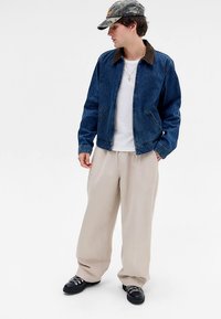 Traper jakna sa smeđim ovratnikom, bijela majica, široke krem hlače i crne cipele. Ležerna odjeća s kapom u kamuflažnom uzorku.