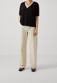 Haut noir à manches courtes avec boutons et col en V, associé à un pantalon ample beige clair et des ballerines noires ornées de clous.