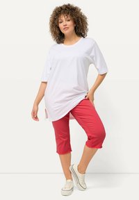 Witte oversized t-shirt met korte mouwen, gecombineerd met rode cropped broek. Model draagt ook witte sneakers. Achtergrond is licht.