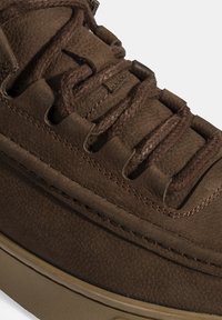 Chaussure en cuir marron avec un dessus texturé, des lacets tressés et des coutures apparentes. La semelle est mate, complétant le design rustique.