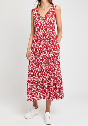 Vrouw met een rood mouwloos midi-jurk met wit bloemenpatroon, witte sneakers en een ketting, staand met handen in de zakken.