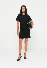 Schwarzes kurzärmeliges Kleid mit Kapuzenärmeln, seitlicher Raffung, Schlitz am Saum, kombiniert mit einer schwarzen strukturierten Clutch und spitzen High Heels.