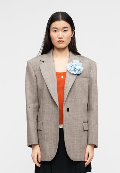 Femme aux longs cheveux noirs portant un blazer beige oversize avec une fleur en tissu bleue, un haut orange et une jupe noire, debout devant un fond blanc.