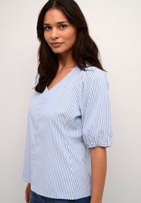 Kék és fehér csíkos blúz V-nyakú, rövid puff ujjakkal és laza szabással. Elöl gombos részlet és sima szövettextúra.