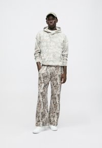 Világosszürke kapucnis pulóver levélmintával, széles szárú nadrággal, hasonló kamuflázs mintázattal kiegészítve, fehér sportcipővel.