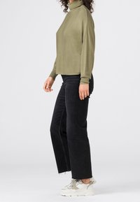 Woman standing sideways wearing an olive green turtleneck sweater, black wide-leg jeans, and white chunky sneakers.