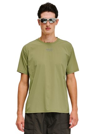 Mężczyzna w oliwkowozielonej koszulce z krótkim rękawem z logo "HALO" oraz czarnych szortach typu cargo, zwrócony przodem, w okularach przeciwsłonecznych z refleksami.