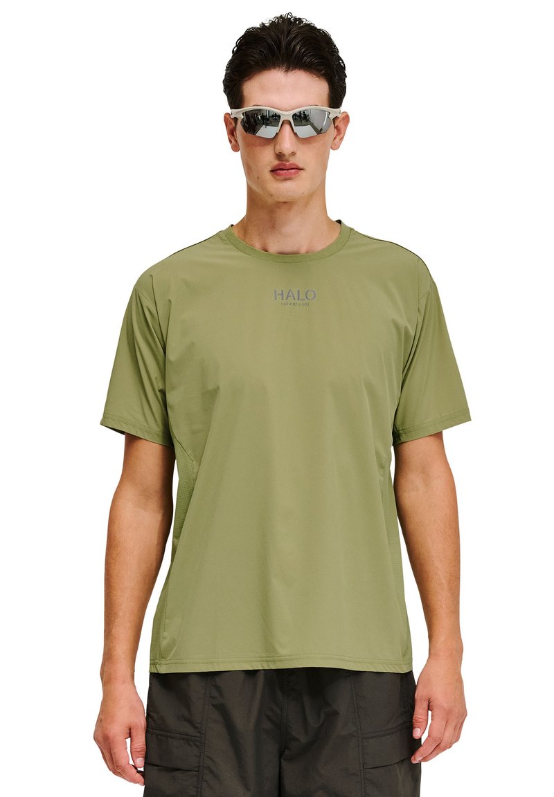 Mężczyzna w oliwkowozielonej koszulce z krótkim rękawem z logo "HALO" oraz czarnych szortach typu cargo, zwrócony przodem, w okularach przeciwsłonecznych z refleksami.
