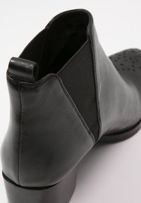 Musta nahkainen nilkkuri, jossa on joustava sivupaneeli, vetotappi takana, matala lohkareunu ja rei'itetty kärkitieto.