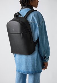 Zaino nero realizzato in materiale testurizzato, con una forma arrotondata, tasca frontale con zip e spallacci imbottiti. Indossato sopra una giacca di jeans.