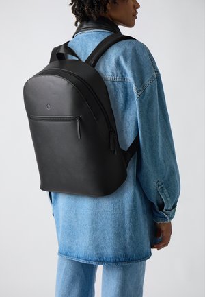 Svart ryggsäck gjord av texturerat material, med rundad form, framficka med dragkedja och vadderade axelremmar. Buren över en denimjacka.
