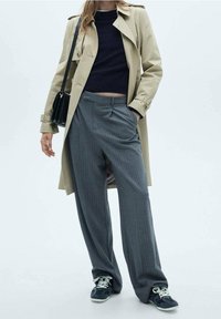 Beige trenchcoat, marineblauwe trui, grijs gestreepte wijde broek en marineblauwe sneakers. Heeft togglesluitingen en een relaxte pasvorm.