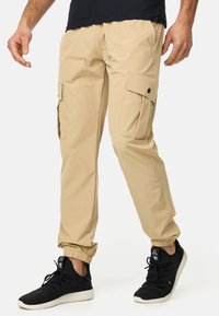 Pantalones cargo beige con puños elásticos, que cuentan con múltiples bolsillos laterales y botones de presión negros, combinados con zapatillas negras.