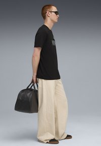 Sort t-shirt med trykt tekst, cremefarvede brede bukser, sorte sneakers og en sort læder rejsetaske. Modellen bærer mørke solbriller.