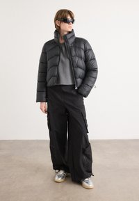 Casaco puffer preto, com textura acolchoada e gola alta. Combinado com calças cargo largas pretas e ténis prateados.