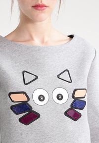 Sudadera gris con un escote amplio que presenta apliques geométricos coloridos, incluyendo círculos y triángulos en acabados metálicos y con purpurina.