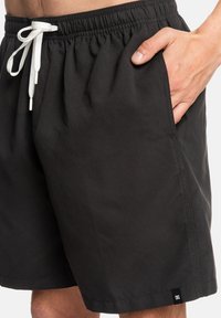 Black shorts with an elastic waistband and white drawstring. Featuring side pockets and a DC logo tag at the hem. Smooth fabric texture.