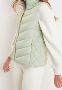 Gilet imbottito verde trapuntato con collo alto, chiusura frontale con zip e tessuto leggero, indossato sopra un maglione bianco a maglia.