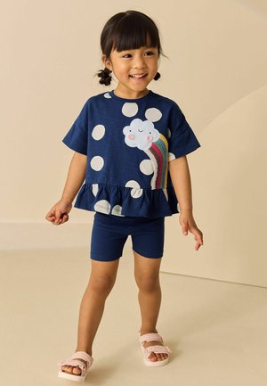 Fiatal lány copfokkal, tengerészkék pöttyös felsővel, melyen felhő és szivárvány minta látható, tengerészkék rövidnadrággal és rózsaszín szandállal, mosolyogva áll.