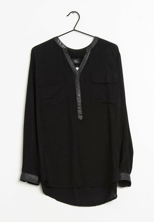 Blouse noire à manches longues avec bordure en cuir synthétique sur le col, la patte de boutonnage et les poignets, avec deux poches poitrine, suspendue sur un cintre noir.