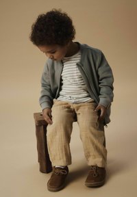 Šedá bomber bunda na zips, pruhované tričko, béžové ľanové nohavice a hnedé semišové topánky. Sedí na drevenej stoličke na neutrálnej pozadí.