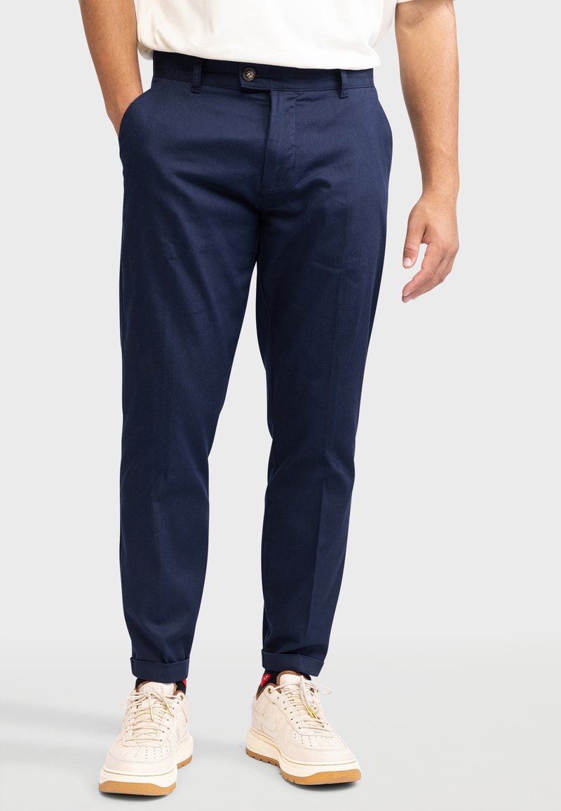 Pantalon marine en tissu léger, avec une coupe slim, des poches avant et une fermeture par bouton. Associé à des baskets beige.