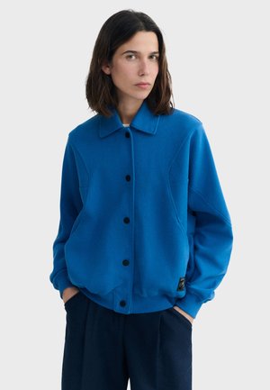 Femme aux cheveux bruns mi-longs portant une veste bleu vif boutonnée, les mains dans les poches d'un pantalon foncé, devant un fond uni.