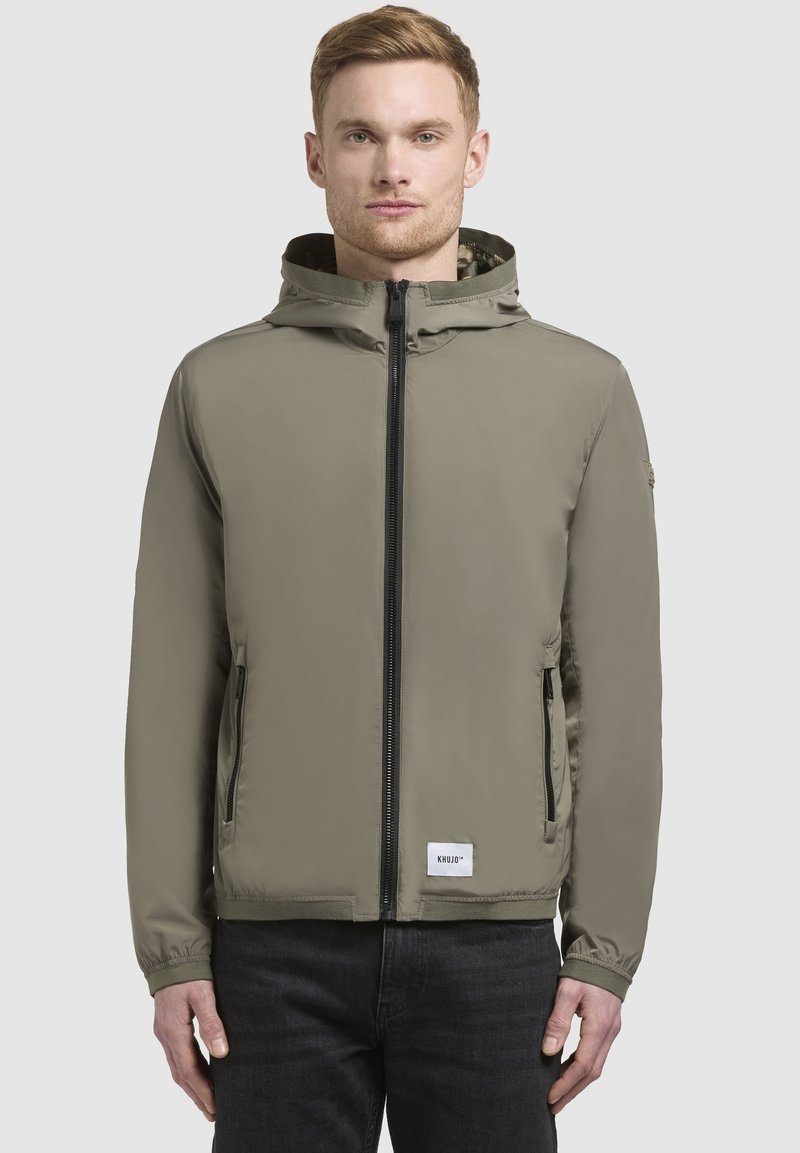 Khakifarbene Kapuzenjacke mit einem Frontreißverschluss, zwei seitlichen Taschen und elastischen Bündchen. Verfügt über ein Logopatch am Saum und eine glatte Textur.