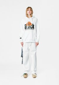 Sudadera blanca con un gran gráfico de "Black" y un perro de dibujos animados, combinada con pantalones blancos holgados. Zapatillas beige con detalles verdes.