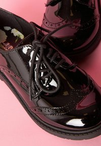 Scarpe in vernice nera con laccio, punta arrotondata e dettagli brogue. Suole testurizzate e lacci neri piatti.