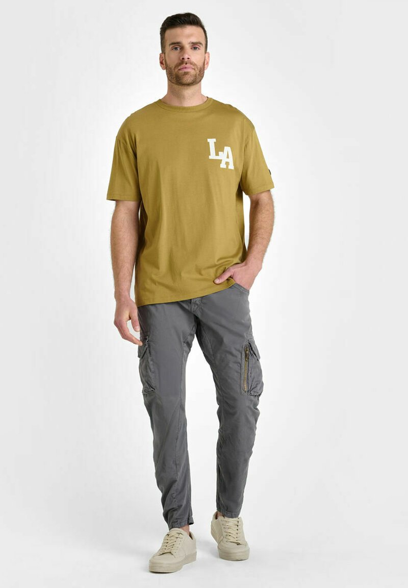 T-shirt vert olive avec impression blanche "LA" sur la poitrine gauche, associé à un pantalon cargo gris avec poches zippées. Baskets de couleur claire.