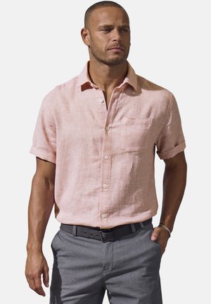 Man wearing a light pink short-sleeve button-up shirt with chest pocket, gray pants, black belt, and bracelet, looking right.
