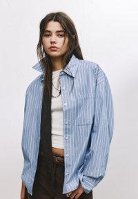 Camicia button-up a righe blu e bianche con colletto ampio, taschino singolo sul petto e vestibilità ampia, indossata sopra una camicia bianca e pantaloni marroni.