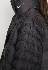 Svart puffjacka med hög krage, med en liten vit Nike-logotyp p å nacken. Tyget har en slät, glänsande yta med kviltade sektioner.