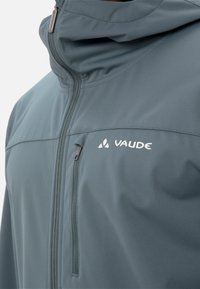 Veste soft-shell de couleur bleu-gris atténué. Elle dispose d'un col haut, d'une poche poitrine zippée et du nom de la marque "VAUDE" imprimé en blanc.