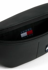 Sacchetto in materiale sintetico nero con chiusura a zip, dotato di un'etichetta "TOMMY JEANS" in blu, bianco e rosso all'interno.