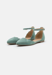 Bailarinas de ante verde con bordes ondulados, punta afilada, correa al tobillo y suela plana, con una textura suave y un diseño minimalista.