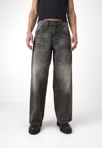 Breda ben, blekta svarta jeans med sliten textur, femficksdesign och nitar i bältet, bärs med svarta skor.