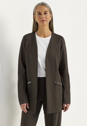 Midaldrende kvinde med gråt hår iført mørkebrun åben blazer, hvid skjorte og matchende bukser, står mod en ensfarvet lys baggrund.