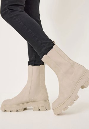 Beige ankle boots with a smooth suede finish, elastic side panels, and chunky rubber soles, paired with black skinny jeans.