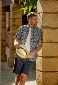 Chemise à manches courtes à fleurs en bleu marine avec des feuilles rouges et vertes, associée à un t-shirt blanc et un short bleu marine ; tenant un chapeau en paille avec un bandeau noir.