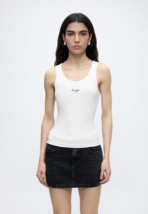 Femme portant un haut blanc sans manches avec le texte "hugo" et une mini-jupe en jean noire, debout devant un fond uni clair.