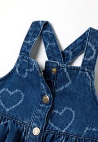 Dunkelblaues Denim-Overallkleid mit Herzmustern, verstellbaren Trägern und silbernen Druckknöpfen mit Herzakzenten.