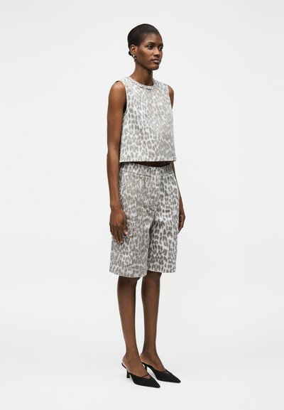 Γυναίκα στέκεται φορώντας γκρι τοπ χωρίς μανίκια με λεοπάρ μοτίβο και αντίστοιχα σορτς μέχρι το γόνατο με μαύρες μυτερές γόβες σε λευκό φόντο.