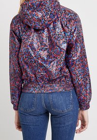 Veste à capuche en matériau brillant avec un motif abstrait coloré en rouge, bleu et violet. Présente un ourlet et des poignets élastiques.