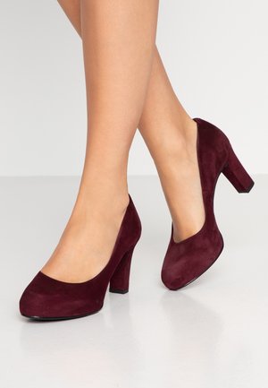 Femme portant des chaussures à talons hauts en suède rouge foncé à bout rond sur un sol gris clair, jambes croisées.