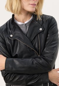 Veste en cuir noir de motard avec un col cranté, quincaillerie argentée et poches zippées, portée sur une chemise grise. Texture lisse et design ajusté.