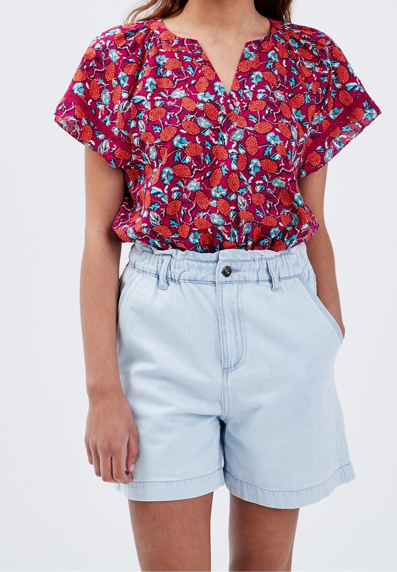 Rote Bluse mit Blumenmuster, weiten Ärmeln, V-Ausschnitt und grünen Akzenten, kombiniert mit hellblauen Jeansshorts mit elastischem Bund.