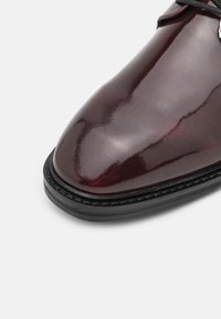 Chaussure habillée en cuir bordeaux, avec une finition brillante, un bout pointu, une semelle noire et des détails cousus le long du rebord.